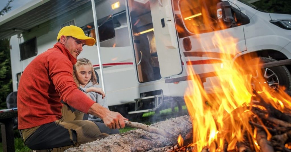 A father and daughter enjoying an open fire while camping next to their RV. 