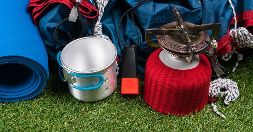 A bunch of camping equipment including a portable stove, some cookware, a bedroll, and other gear packed in bags.